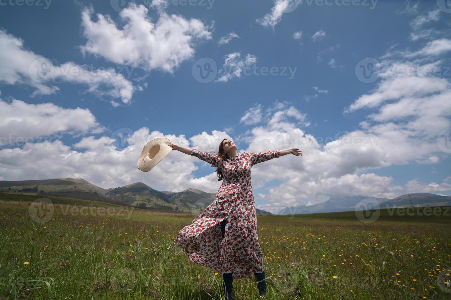 linda mulher hispânica em um vestido florido e chapéu levantando os braços e a cabeça como um sinal de felicidade no meio de um campo foto