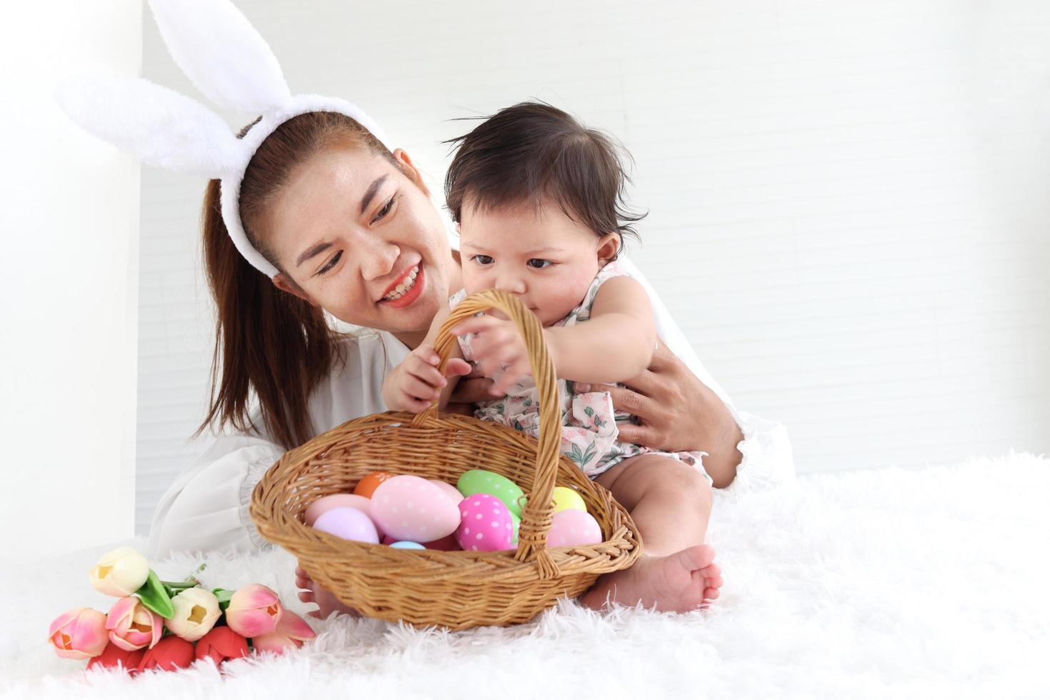 feliz sorrindo adorável menina de seis meses brincando com cesta de vime de ovos de páscoa coloridos no abraço da mãe braços, mãe com cabeça de orelhas de coelho segurando seu doce filhinha. foto