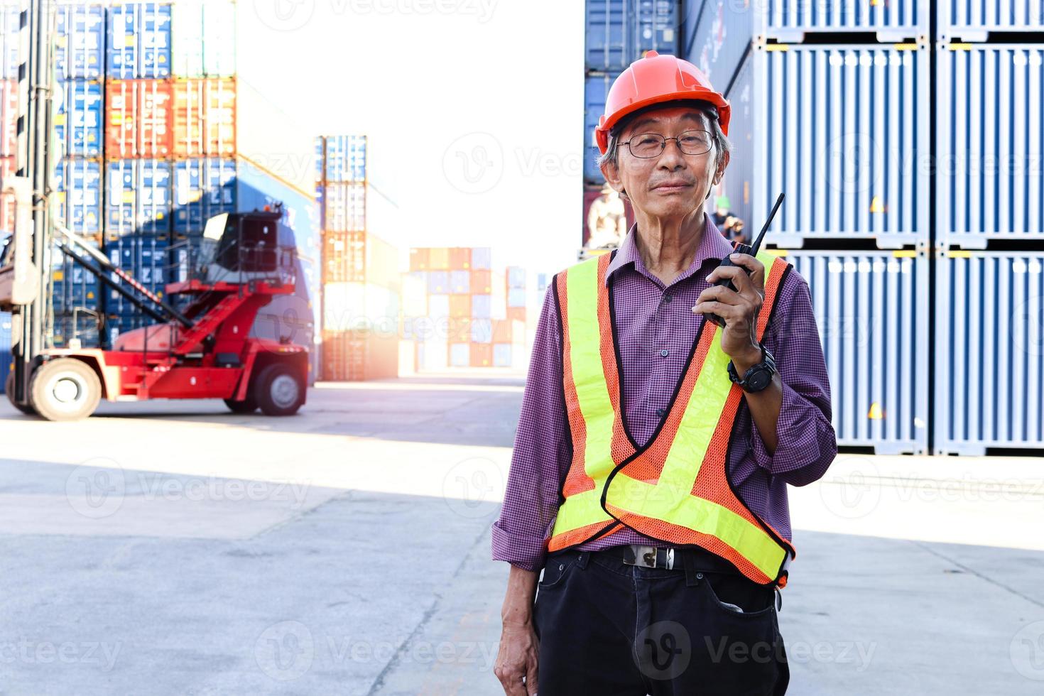 um engenheiro trabalhador asiático idoso sênior usando colete de segurança e capacete em pé e segurando walkies talkie de rádio no pátio de contêineres de carga logística. pessoas idosas no conceito de local de trabalho. foto