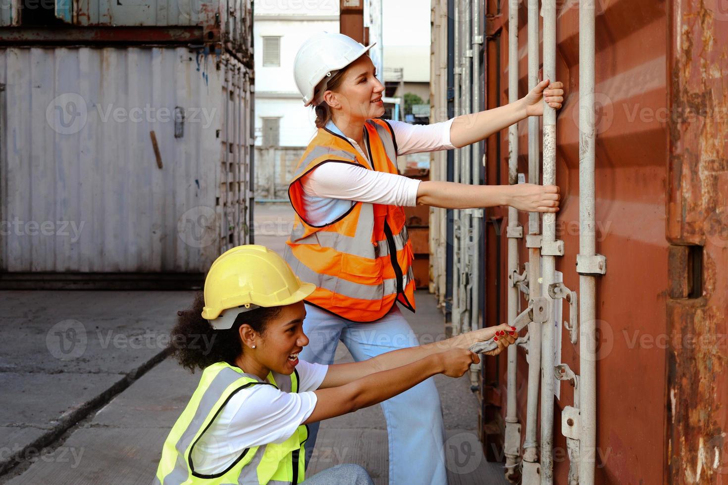 duas belas mulheres engenheiras trabalhadoras usam colete de segurança e capacete, tentam abrir a porta do contêiner no pátio de carga logística. garotas afro-americanas e caucasianas trabalham juntas no mesmo local de trabalho. foto