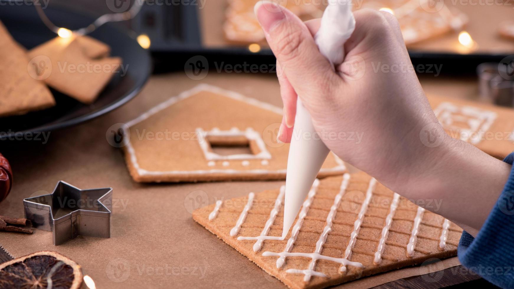 mulher está decorando a casa de biscoitos de gengibre com cobertura de glacê branco no fundo da mesa de madeira, papel manteiga na cozinha, close-up, macro. foto