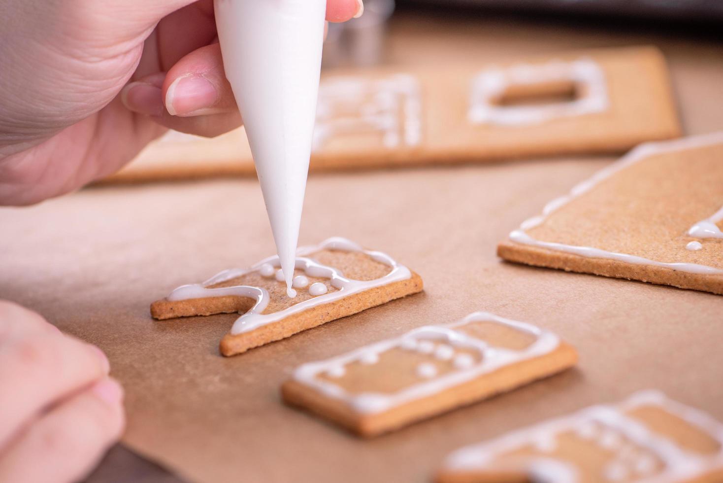 mulher está decorando a casa de biscoitos de gengibre com cobertura de glacê branco no fundo da mesa de madeira, papel manteiga na cozinha, close-up, macro. foto