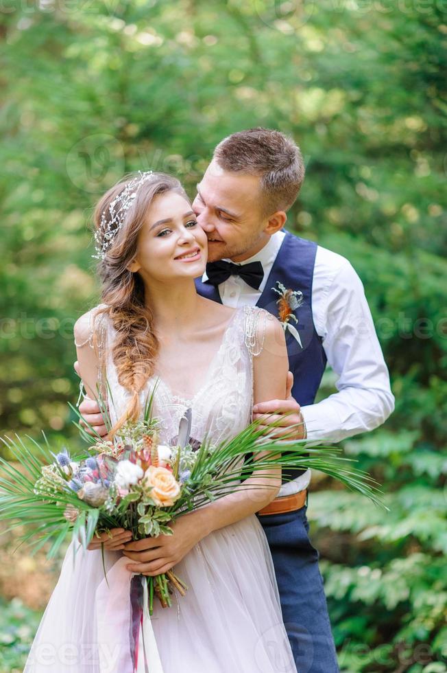 um atraente casal recém-casado, um momento feliz e alegre. um homem e uma mulher se barbeiam e se beijam com roupas de férias. cerimônia de casamento de estilo boêmio na floresta ao ar livre. foto