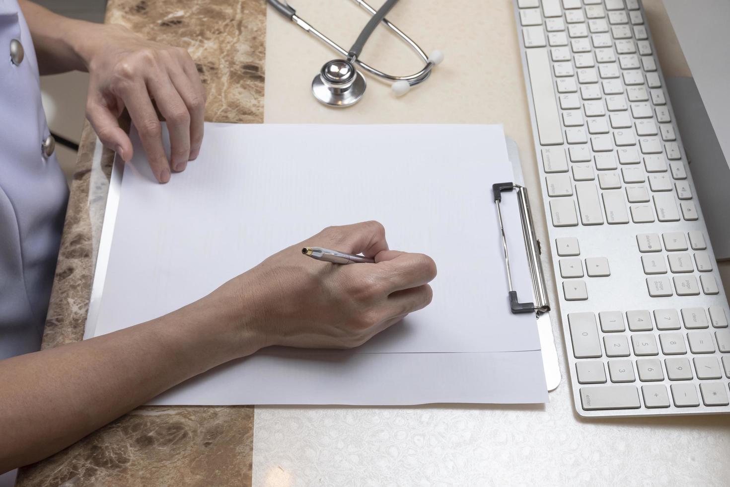 médica ou enfermeira asiática está escrevendo algo na área de transferência com estetoscópio e parte do teclado do computador em cima da mesa na sala do consultório médico foto