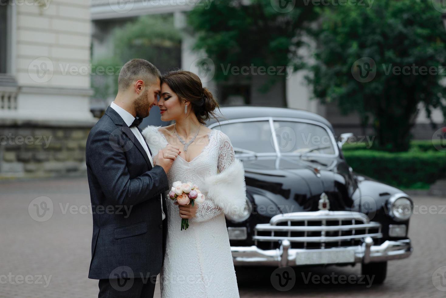 casal de noivos de luxo feliz beijando e abraçando perto de retrô com carro de buquê no outono foto