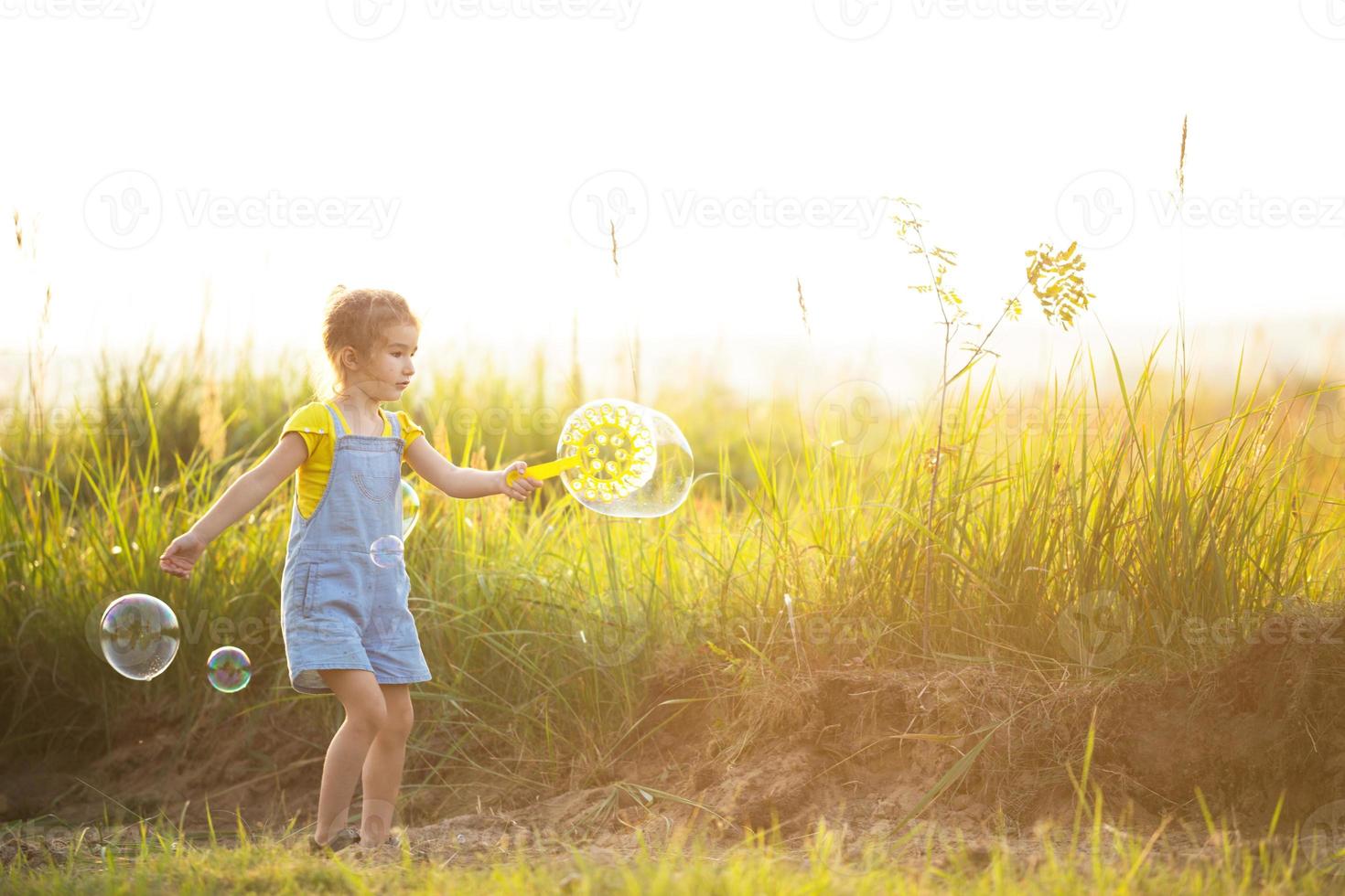 uma garota em um macacão jeans sopra bolhas de sabão no verão em um campo ao pôr do sol. dia internacional da criança, criança feliz, atividades ao ar livre. fundo de verão. estilo de vida saudável e ecológico foto