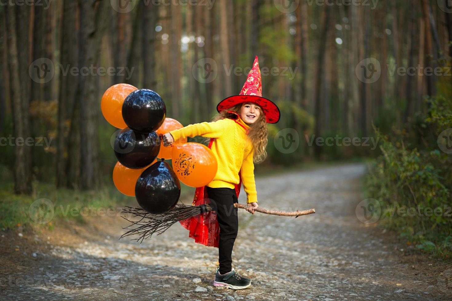 uma garota com uma fantasia de bruxa e chapéu em uma vassoura com balões laranja e pretos está brincando na floresta de outono, indo para uma festa de halloween foto