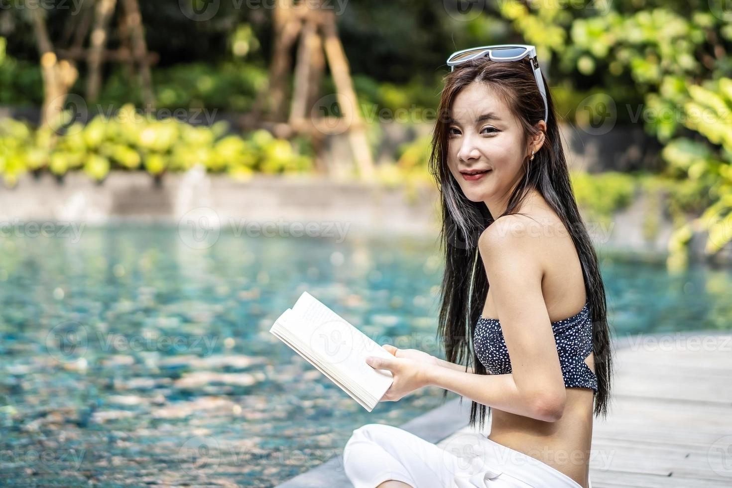 bela jovem asiática senta-se na beira da piscina ao ar livre. ela lendo livro enquanto relaxa perto da piscina foto