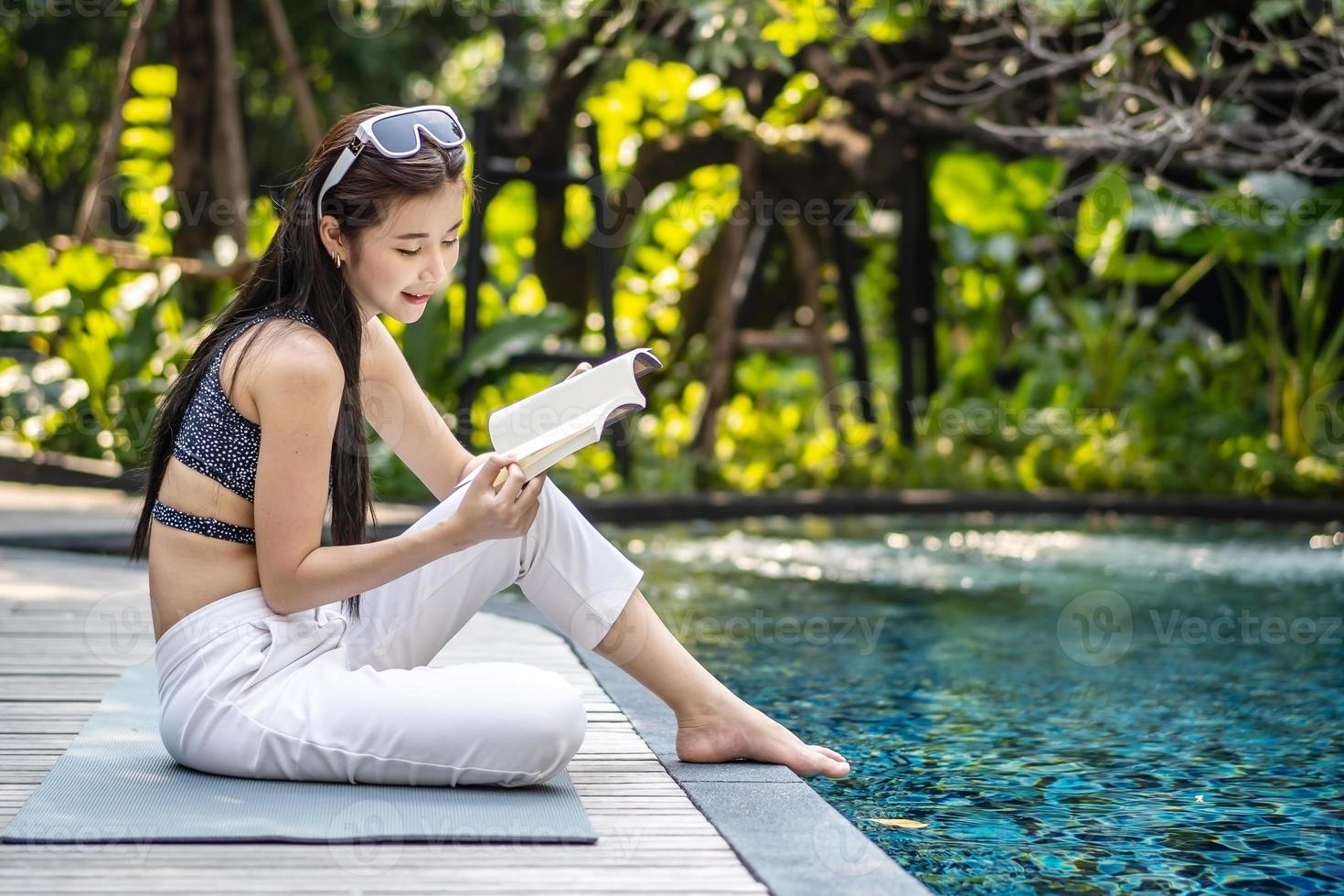 bela jovem asiática senta-se na beira da piscina ao ar livre. ela lendo livro enquanto relaxa perto da piscina foto