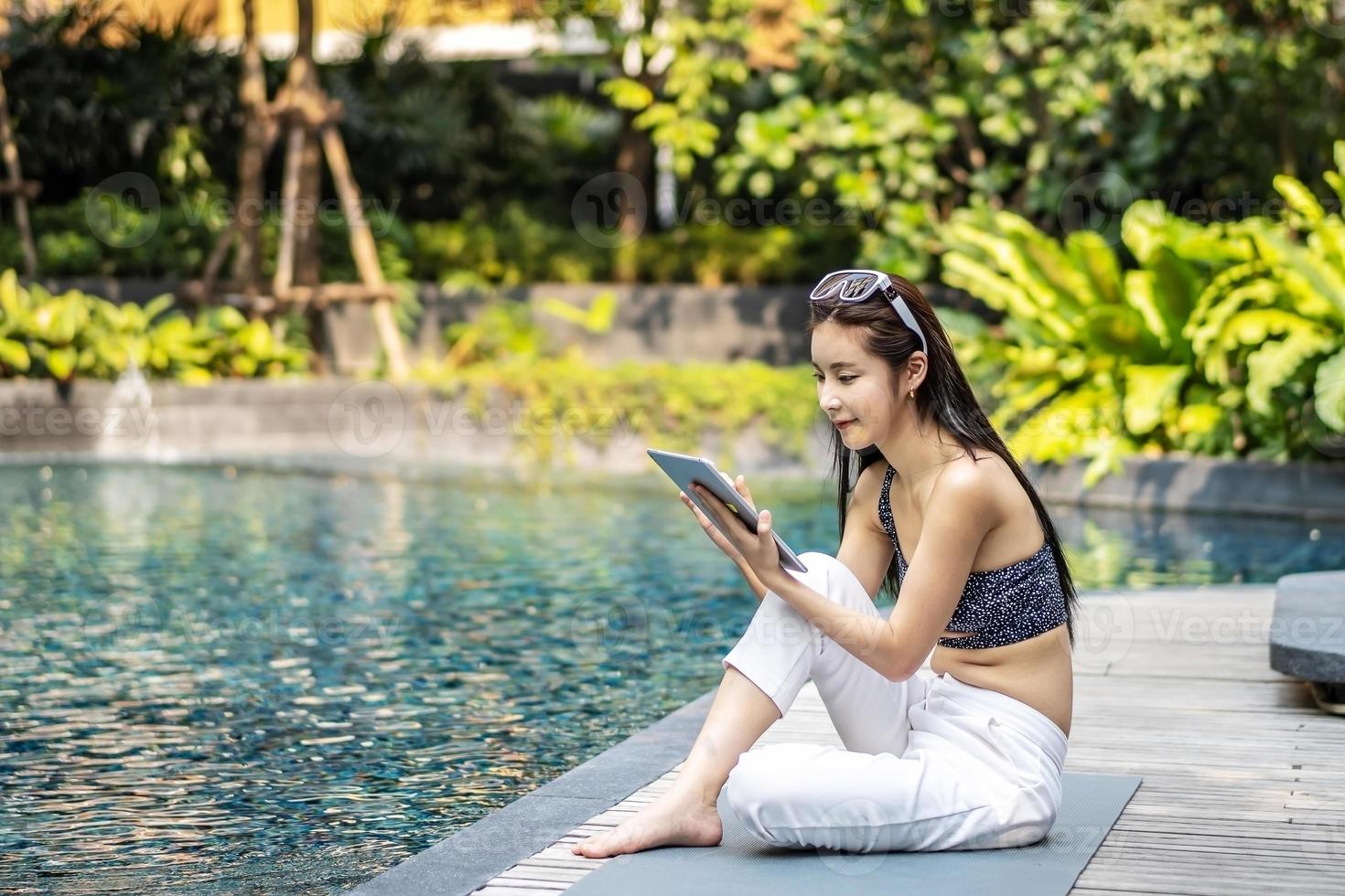 linda mulher asiática relaxante com o uso de computador tablet digital ao ar livre à beira da piscina foto