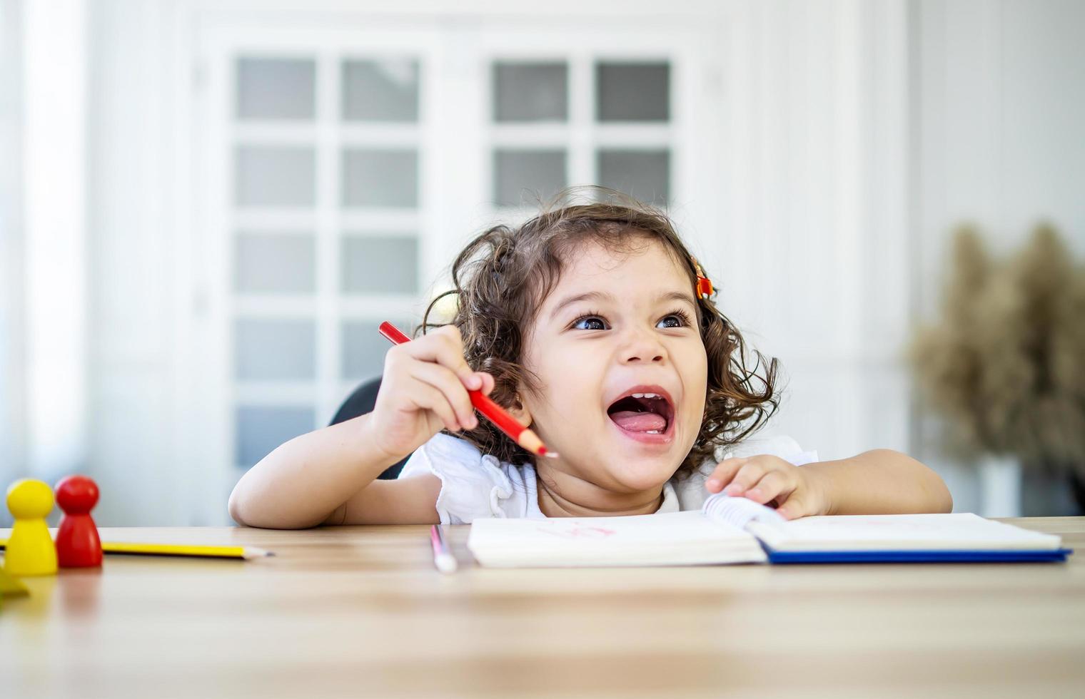 menina engraçada sente-se na mesa em casa fazendo lição de casa, lendo, escrevendo e pintando. crianças pintam. crianças desenham. pré-escolar com livros em casa. pré-escolares aprendem a escrever e ler. criança criativa foto