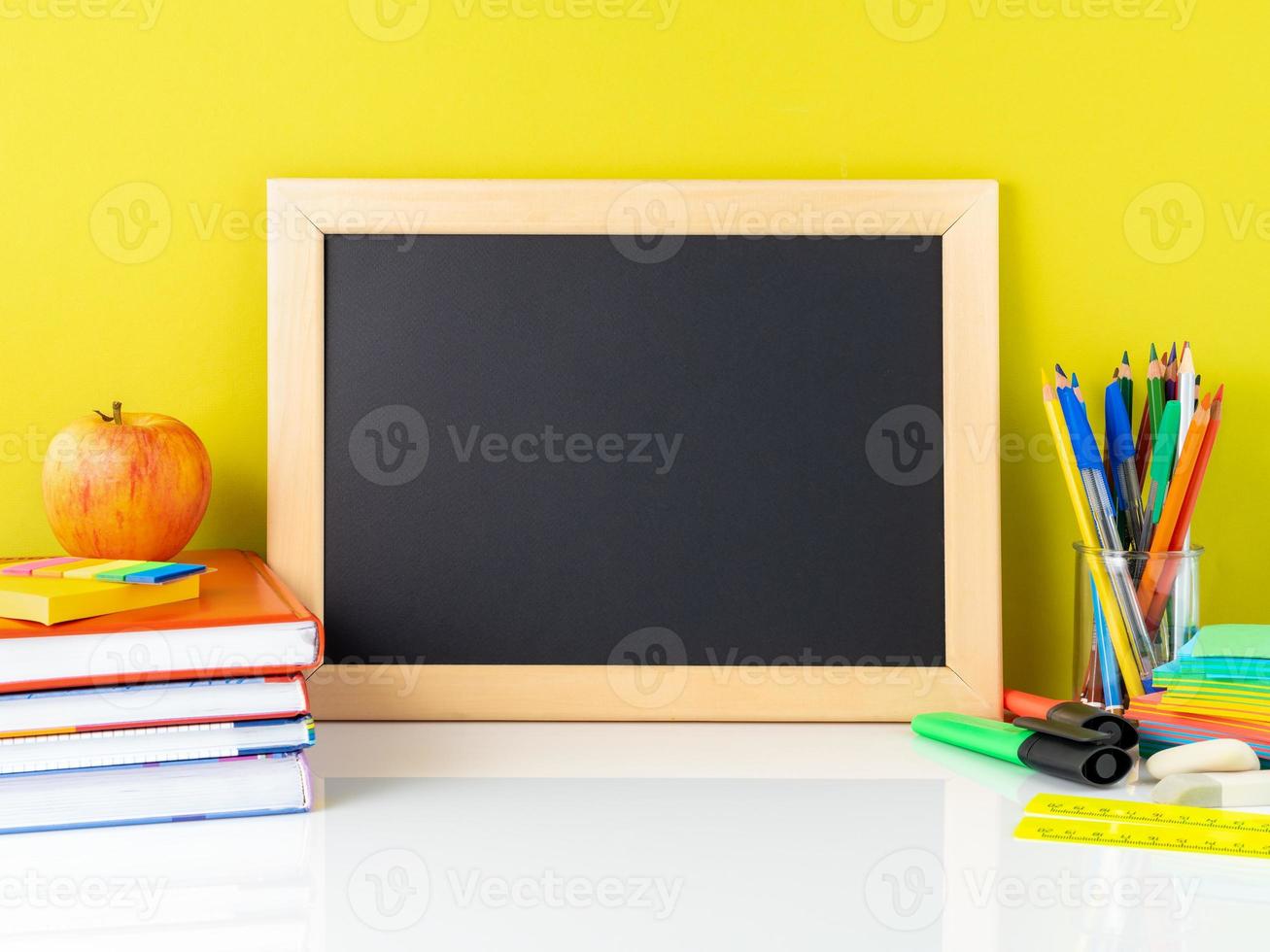 quadro-negro, maçã e material escolar na mesa branca pela parede amarela. vista lateral, espaço vazio. de volta ao conceito de escola. foto