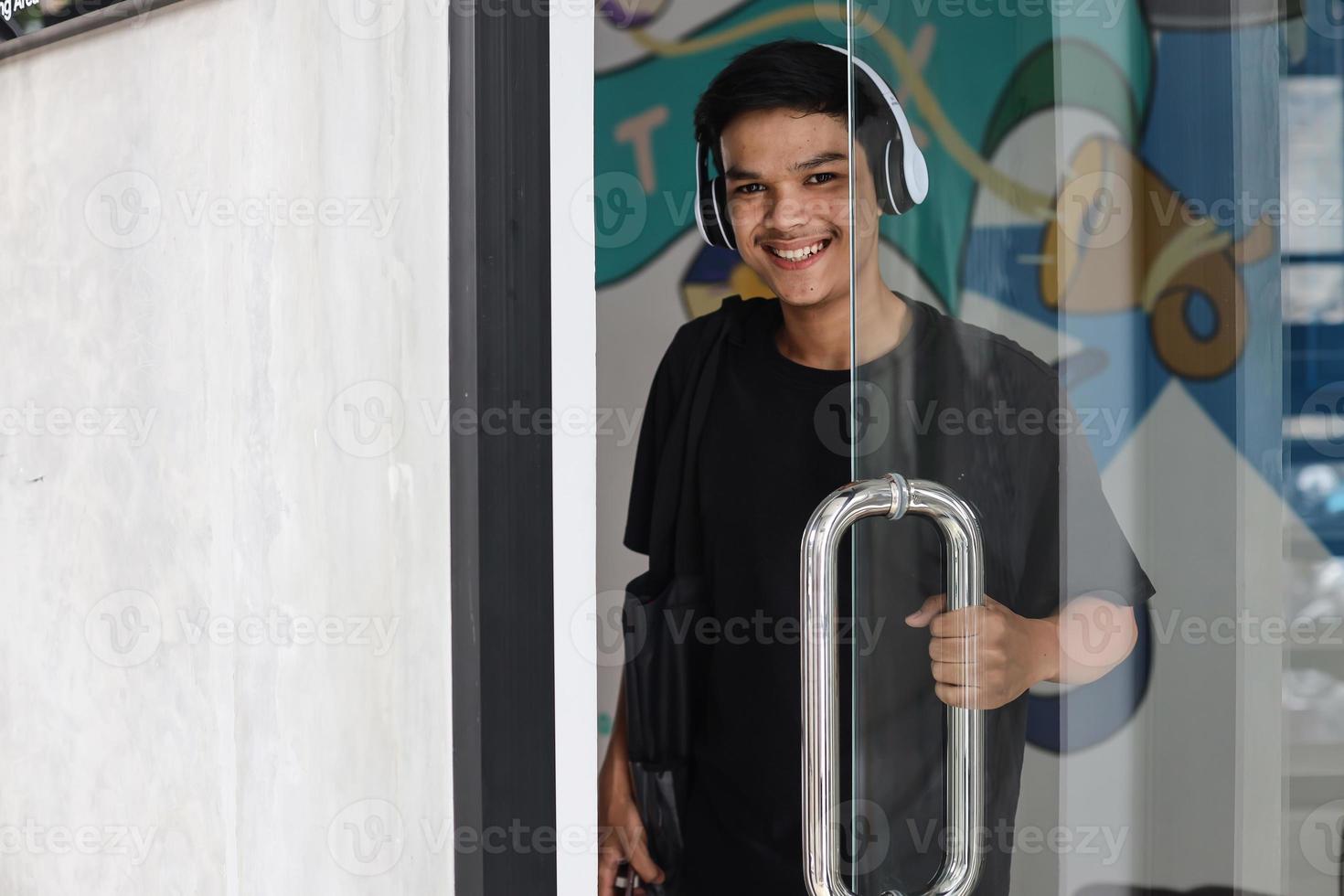 feliz jovem asiático usando fones de ouvido sem fio sorrindo para a câmera ao abrir a porta de vidro foto