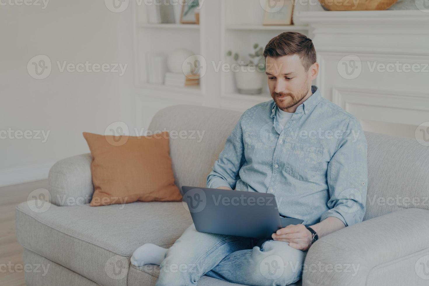 atencioso jovem bonito em roupas casuais usando laptop enquanto está sentado no sofá foto