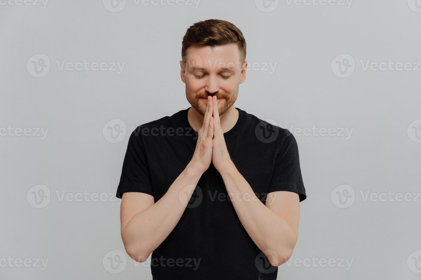 jovem feliz em camiseta preta com os olhos fechados, mantendo as palmas das mãos pressionadas e sonhando com algo foto