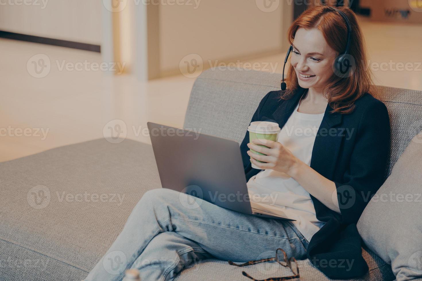 agente de call center feminino jovem sorridente no fone de ouvido sem fio e laptop sentado no sofá no café foto