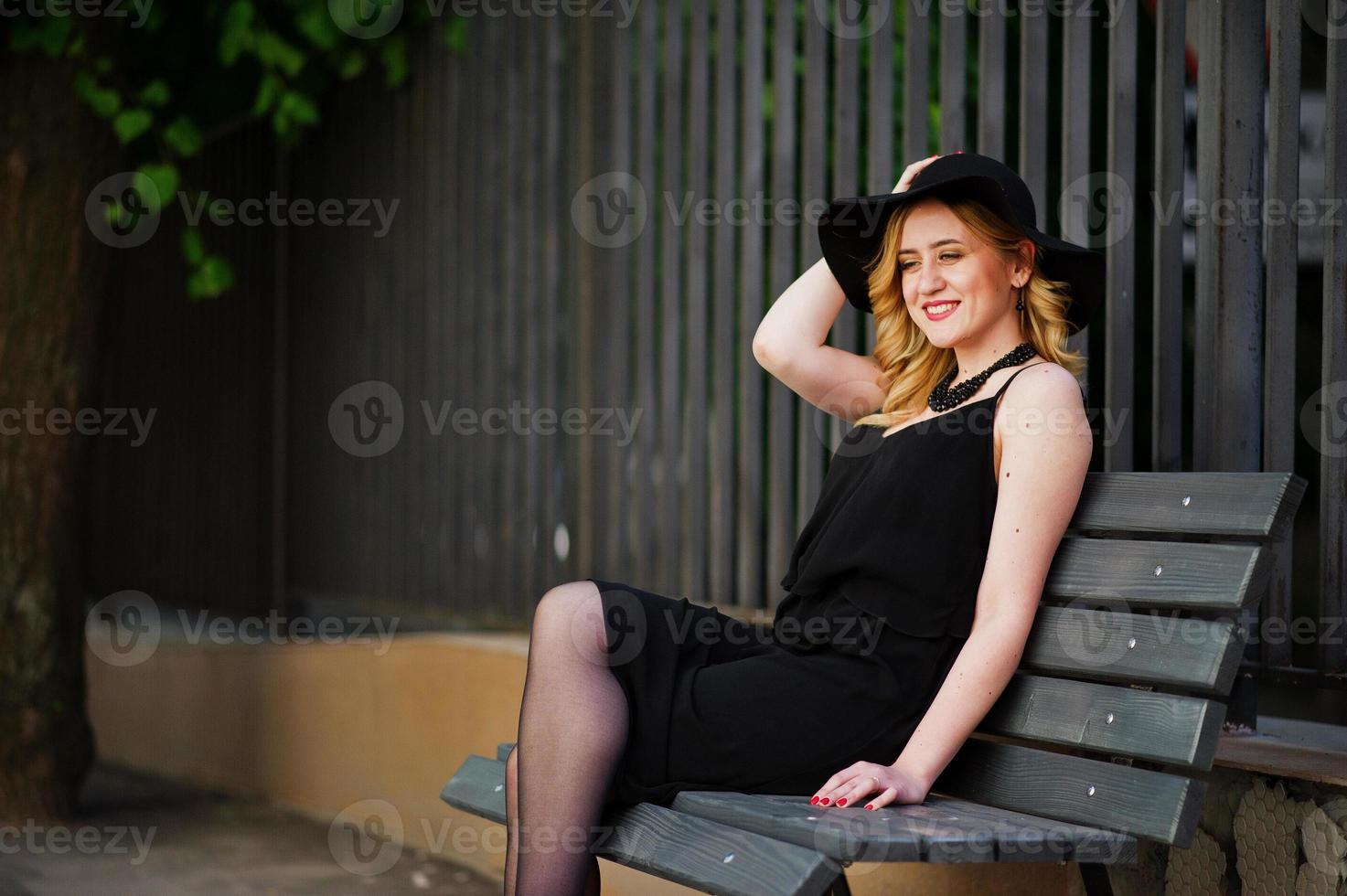 mulher loira de vestido preto, colares e chapéu sentado no banco. foto