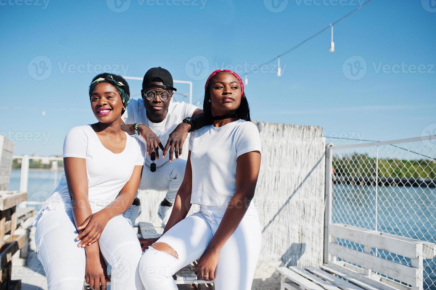 três amigos afro-americanos elegantes, usam roupas brancas no cais na praia. moda de rua de jovens negros. homem negro com duas garotas africanas. foto