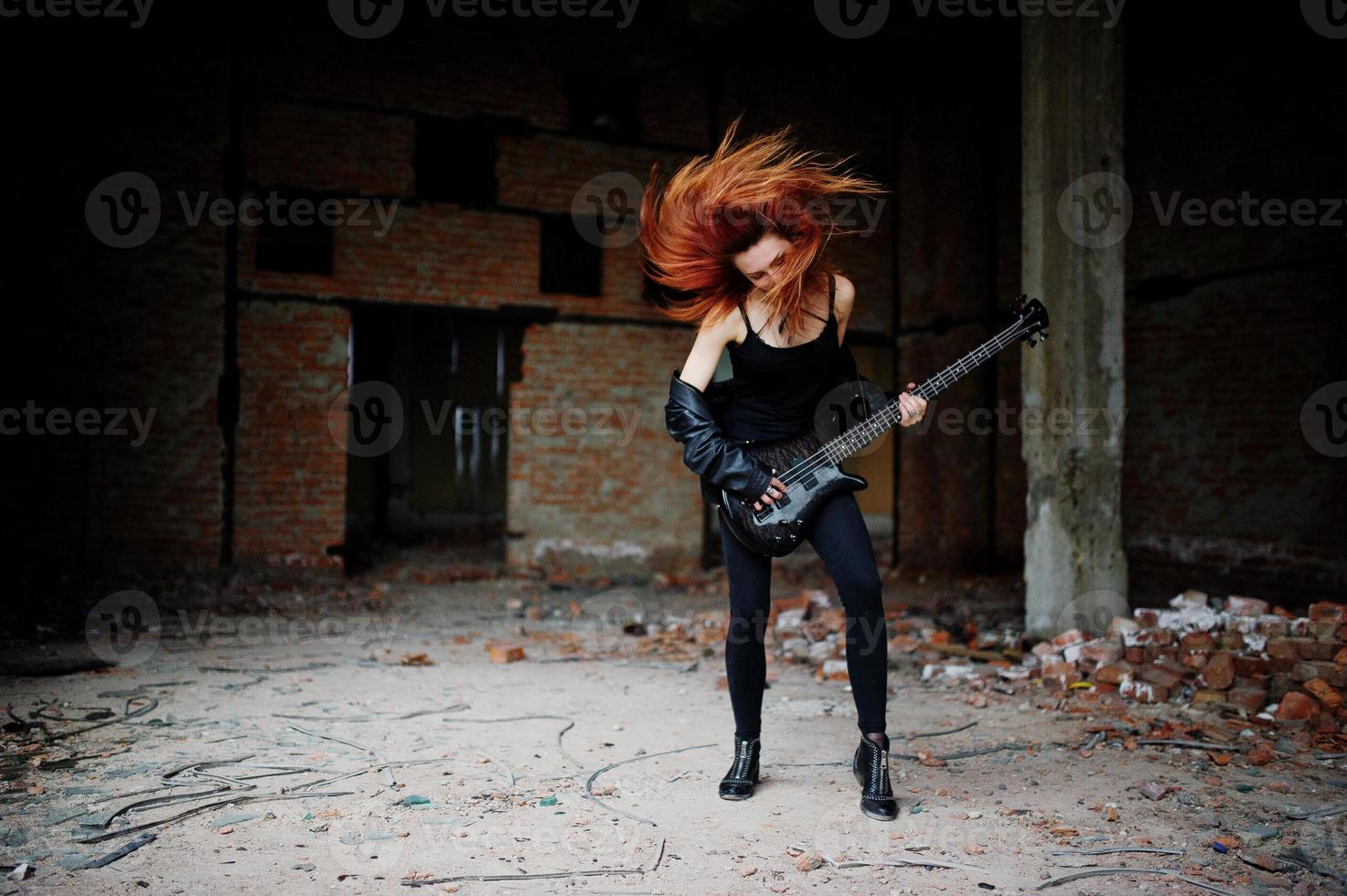 garota punk de cabelo vermelho usa preto com guitarra baixo em lugar abandonado. retrato de músico de mulher gótica. foto