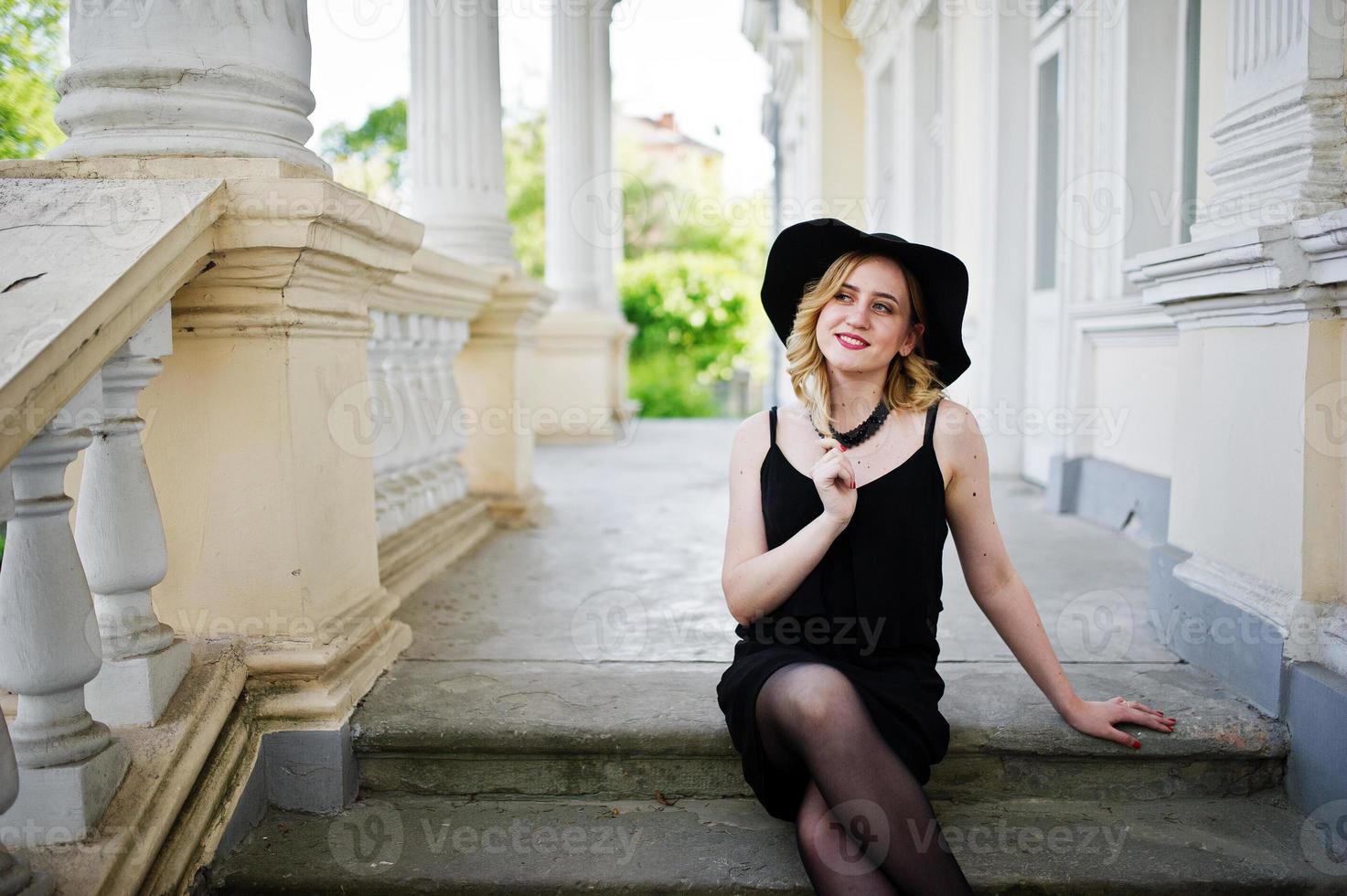 mulher loira de vestido preto, colares e chapéu contra casa vintage. foto