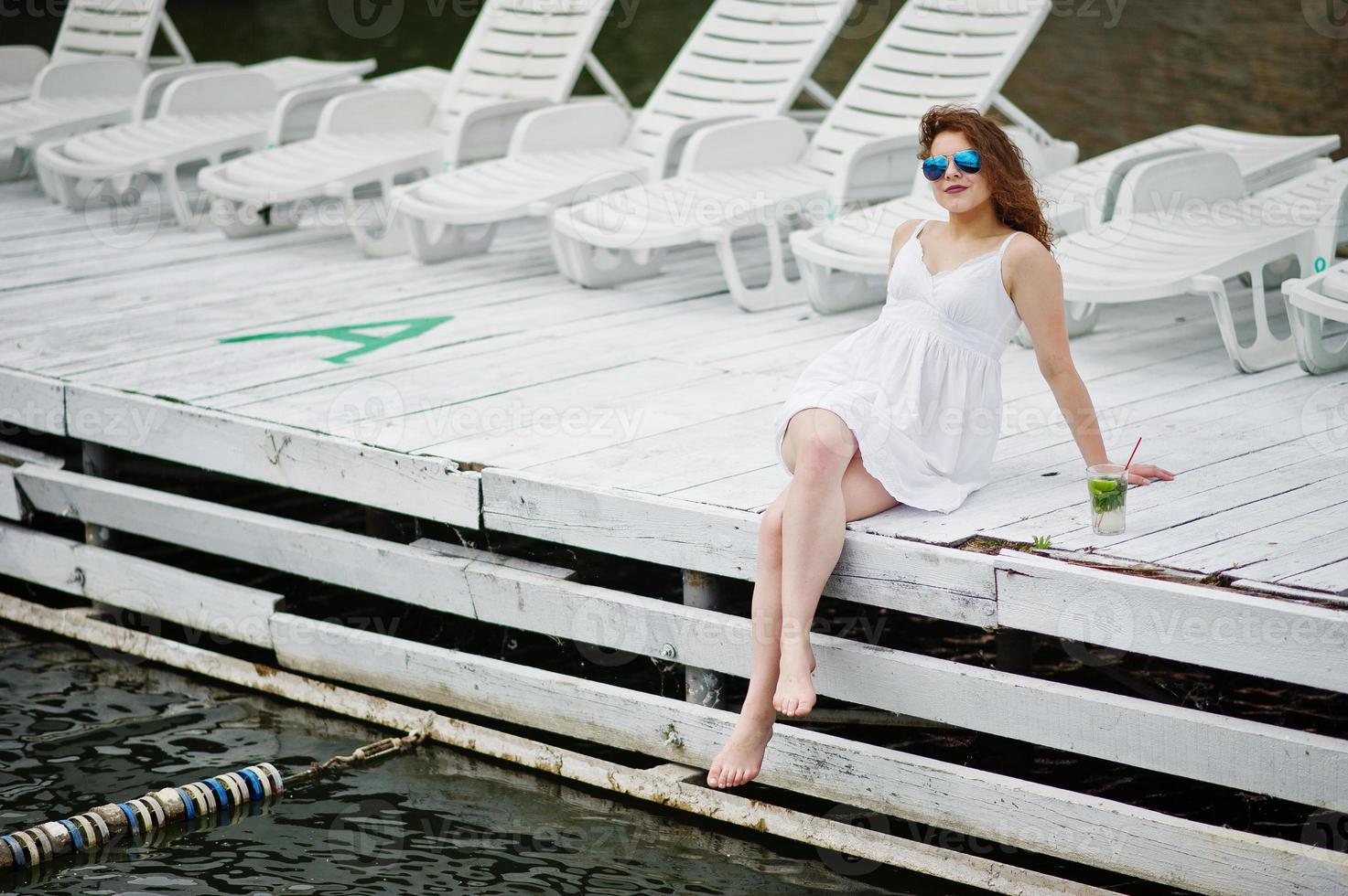 retrato de uma jovem fantástica com óculos de sol e sua bebida sentado no porto de madeira branco ao lado da água. foto
