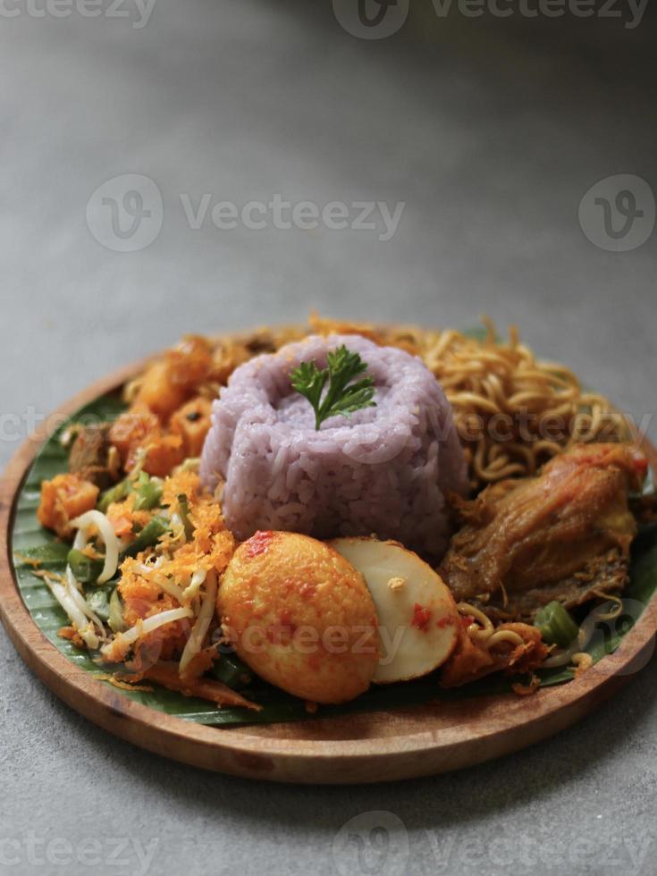 vista de alto ângulo de arroz roxo com condimentos tradicionais indonésios, como ovo picante, macarrão frito, frango picante, batata frita, na placa de madeira e mesa de cor cinza irregular. foto