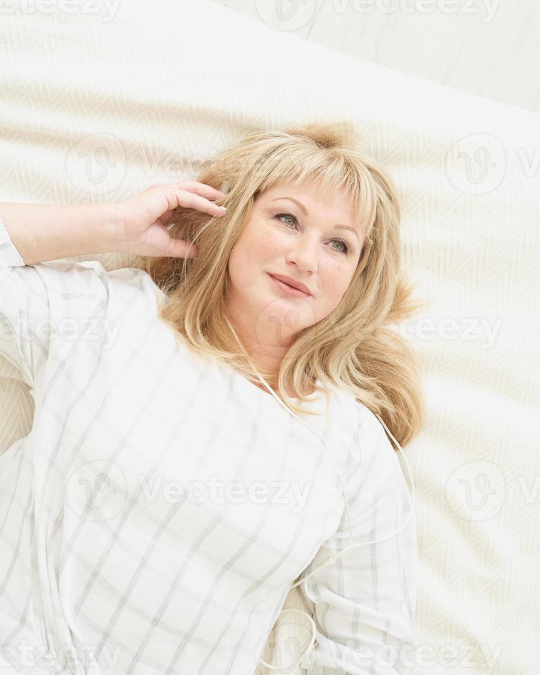 mulher madura com fones de ouvido relaxando em casa, deitada na cama e ouvindo música. foto