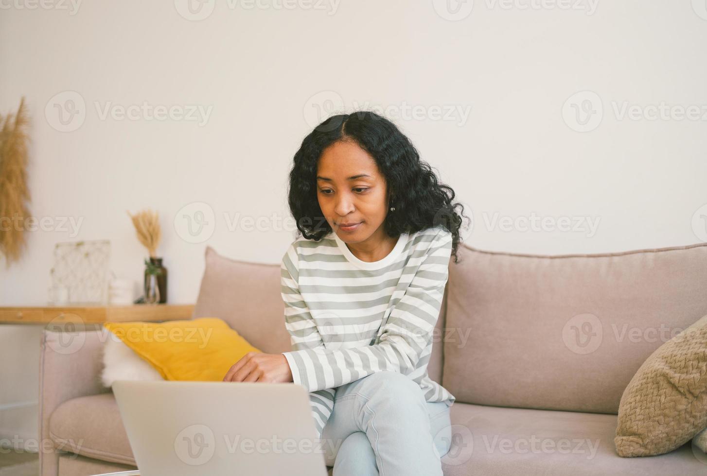 laptop de navegação feminino afro-americano enquanto está sentado no sofá. usando dispositivo digital para trabalhar, estudar foto