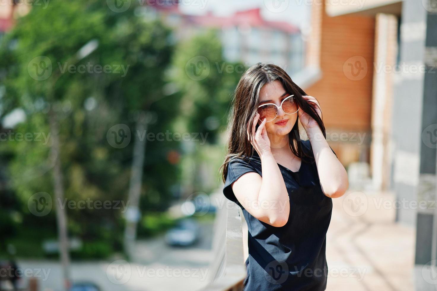 menina morena de vestido preto, óculos de sol posando na rua da cidade. foto