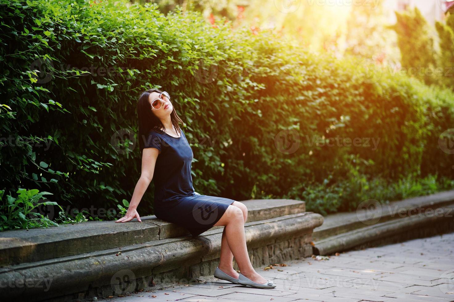 menina de negócios morena de vestido preto em óculos de sol, sentado na fronteira contra arbustos, posando na rua da cidade. foto