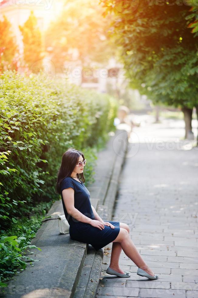 menina de negócios morena de vestido preto em óculos de sol, sentado na fronteira contra arbustos, posando na rua da cidade. foto