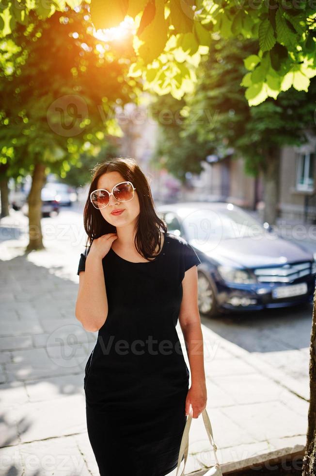 menina de negócios morena de vestido preto em óculos de sol com bolsa na mão posando na rua da cidade, carro de fundo na estrada. foto