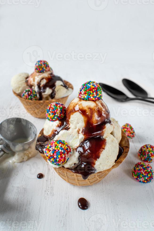 bolas de sorvete de baunilha com pedaços de doces coloridos e calda de chocolate na tigela de cone de waffle em fundo branco rústico foto