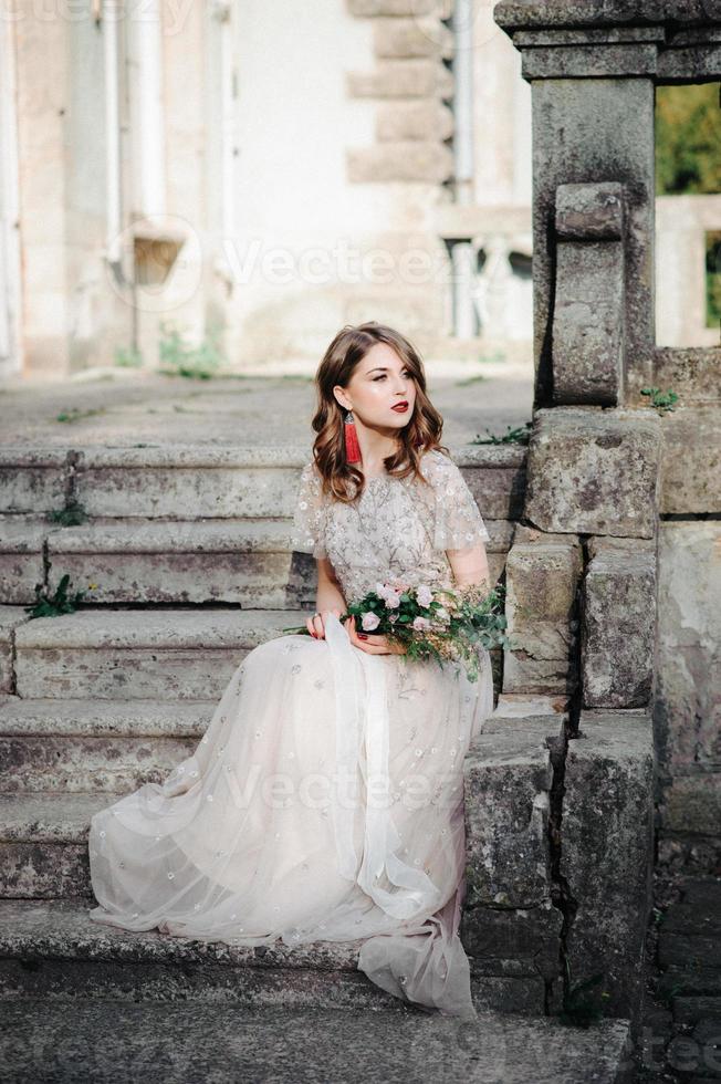 modelo bride.young com pele perfeita e maquiagem, fundo branco. linda noiva na escada de fundo branco. uma mulher em um longo vestido branco está sentada na escada. foto