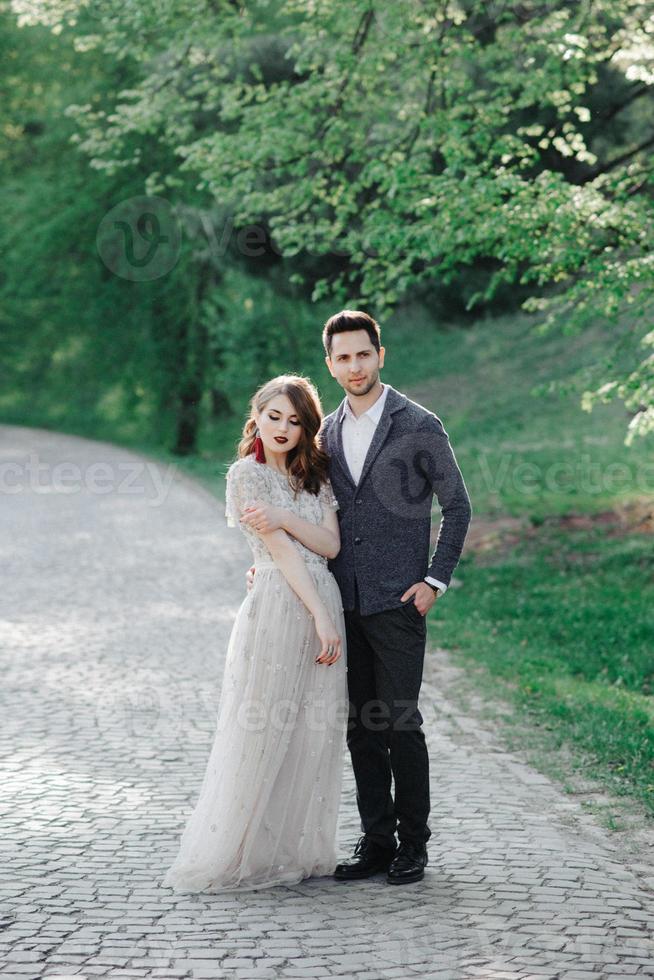 casal em trajes de casamento com um buquê de flores e vegetação está nas mãos contra o pano de fundo do campo ao pôr do sol, a noiva e o noivo foto