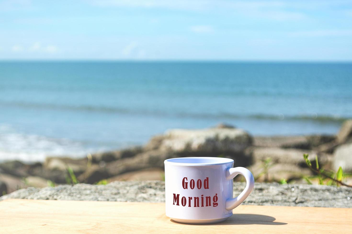 texto de bom dia em uma xícara de café branco com fundo de praia desfocado foto
