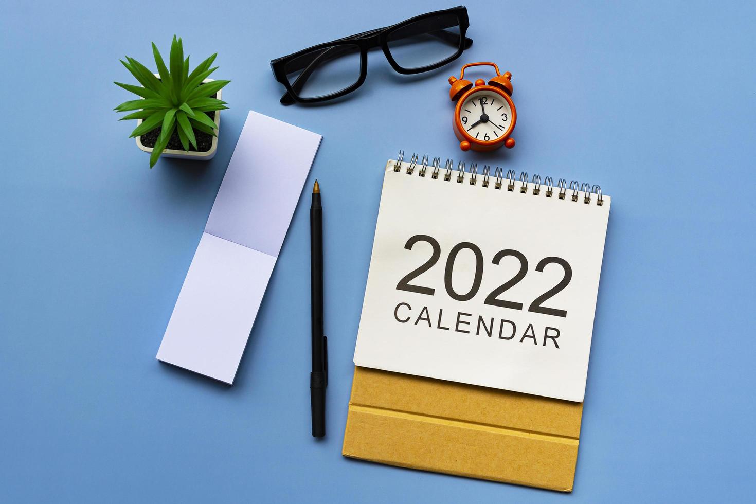 bloco de notas com calendário 2022, óculos, caneta, vaso de plantas e despertador. foto