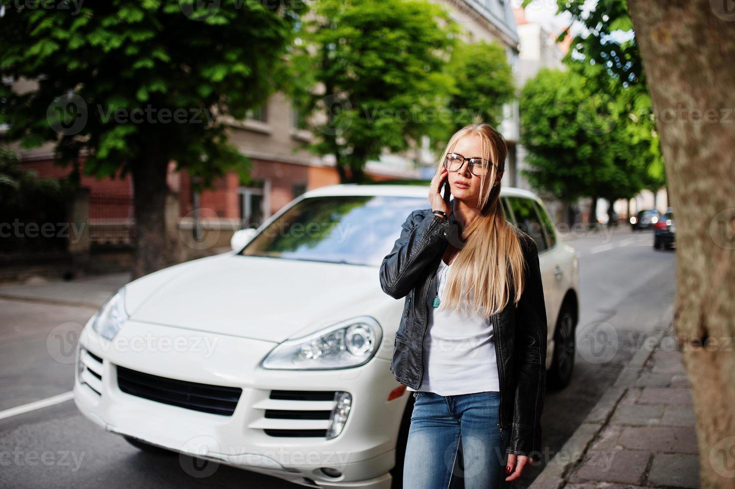 mulher loira elegante usa jeans, óculos e jaqueta de couro com telefone celular, contra carro de luxo. retrato de modelo urbano de moda. foto