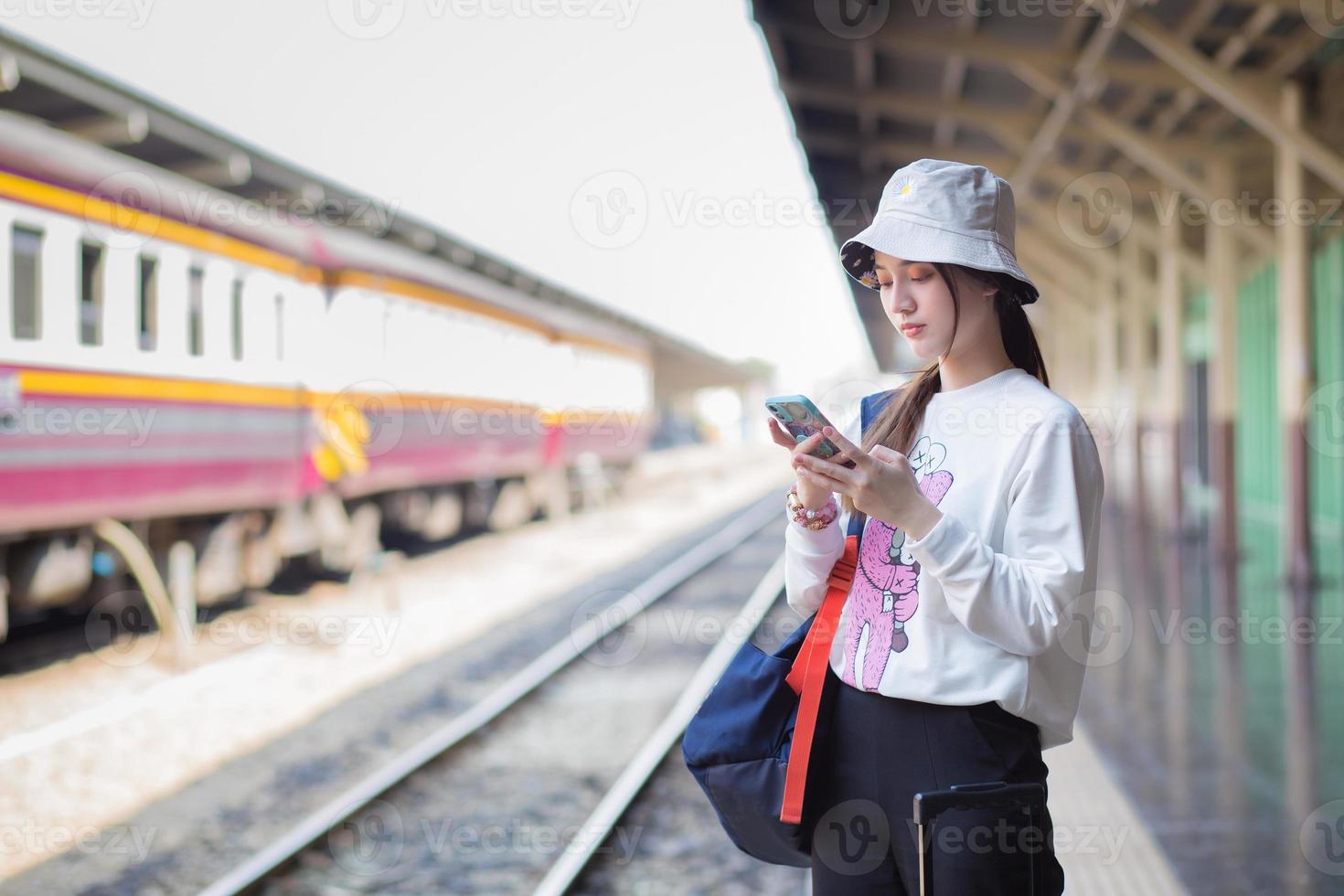 uma linda mulher asiática segurando uma bolsa e um smartphone em busca de informações de viagem. enquanto espera o trem na plataforma foto