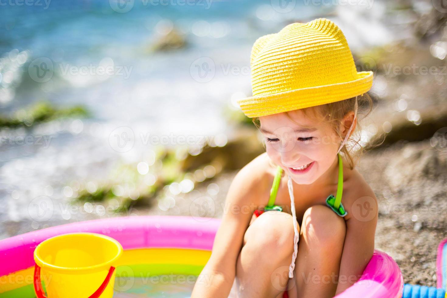 garota de chapéu de palha amarelo brinca com o vento, água e um dispensador de água em uma piscina inflável na praia. produtos indeléveis para proteger a pele das crianças do sol, queimaduras solares. resort no mar. foto
