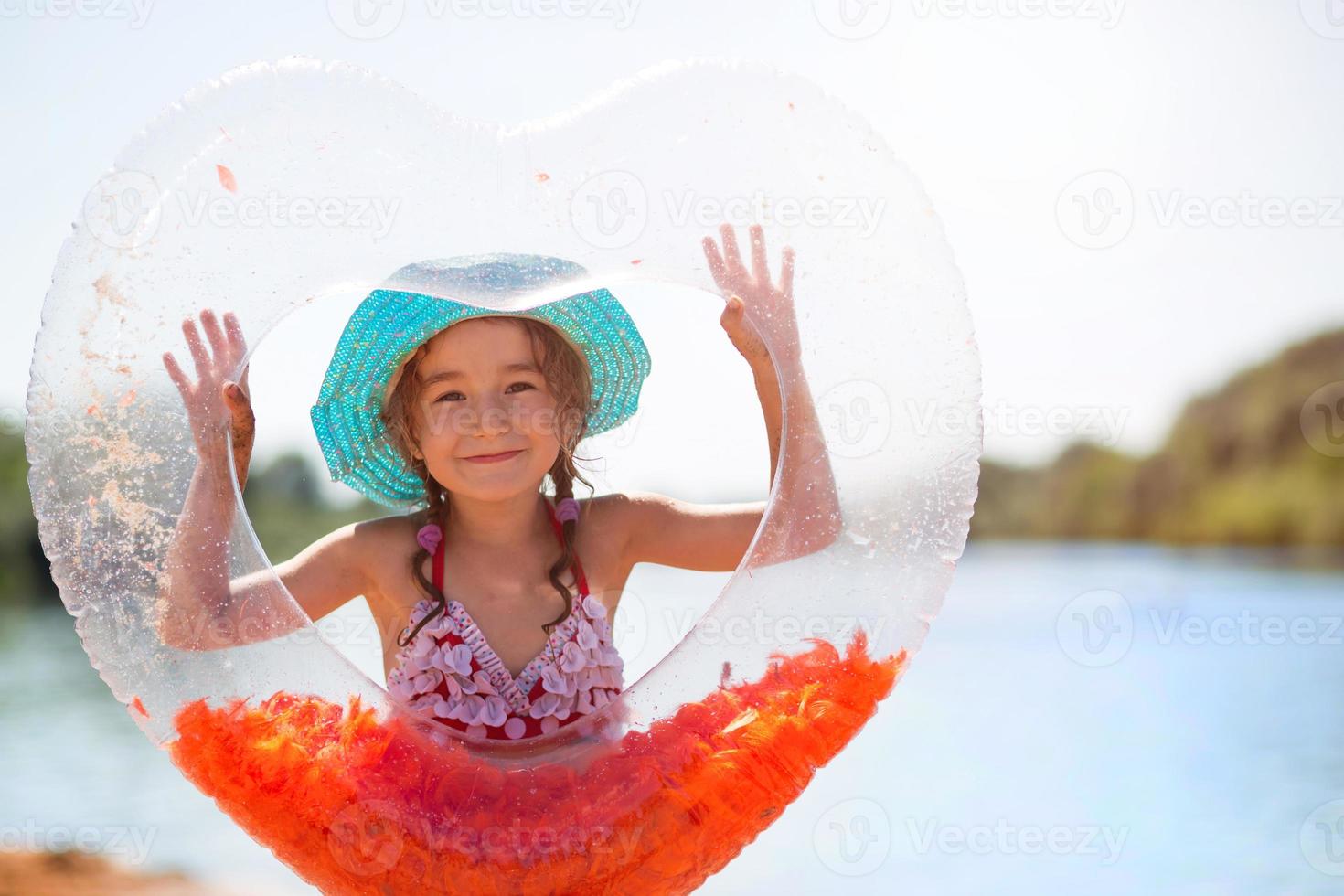 menina de chapéu fica na margem do rio com um círculo inflável transparente em forma de coração com penas laranja dentro. férias na praia, natação, bronzeamento, protetores solares. foto