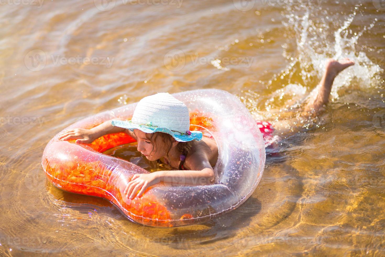 garota de chapéu nadando no rio com um círculo inflável transparente em forma de coração com penas laranja dentro. o mar com fundo arenoso. férias na praia, natação, bronzeamento, protetores solares. foto