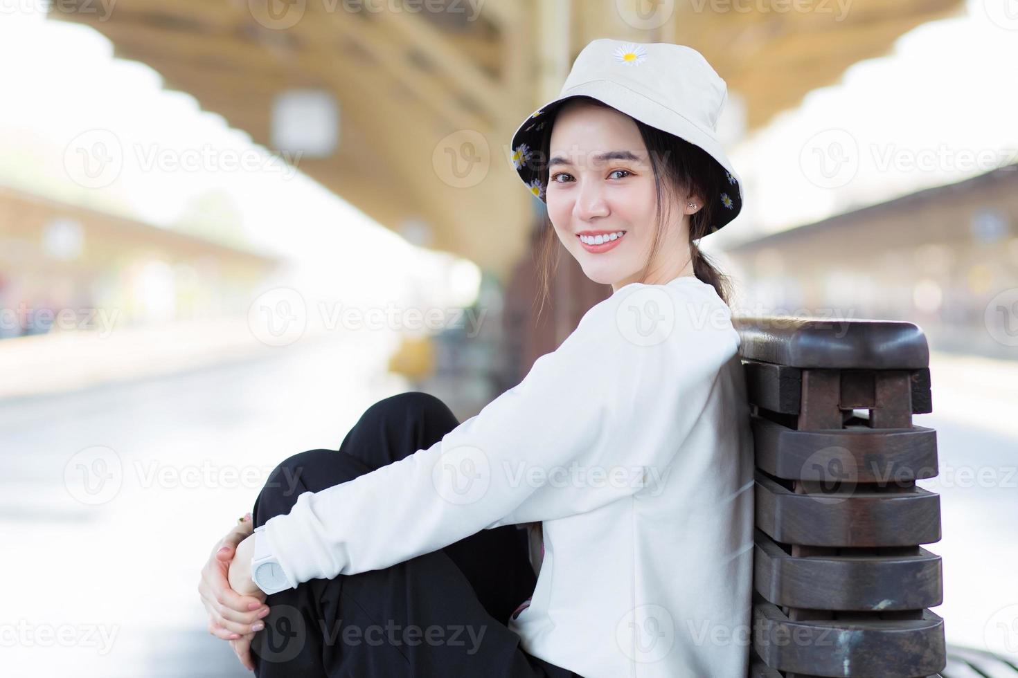 mulher bonita asiática na camisa branca de manga comprida e chapéu sente-se na estação de trem para esperar o trem chegou. foto