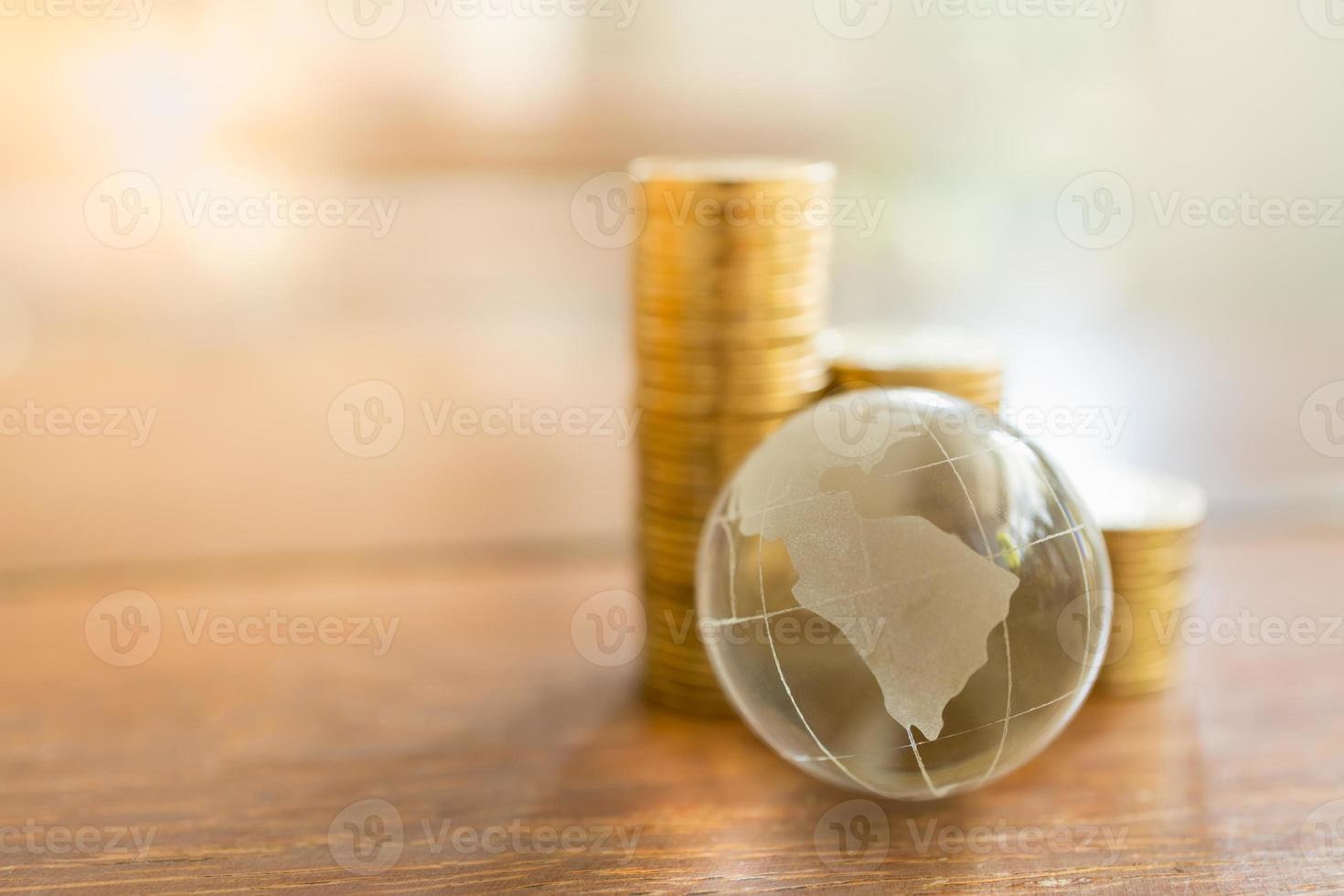 negócios globais e conceito financeiro. close-up da mini bola do mundo de cristal claro com pilha de moedas de ouro na mesa de madeira e copie o espaço. foto