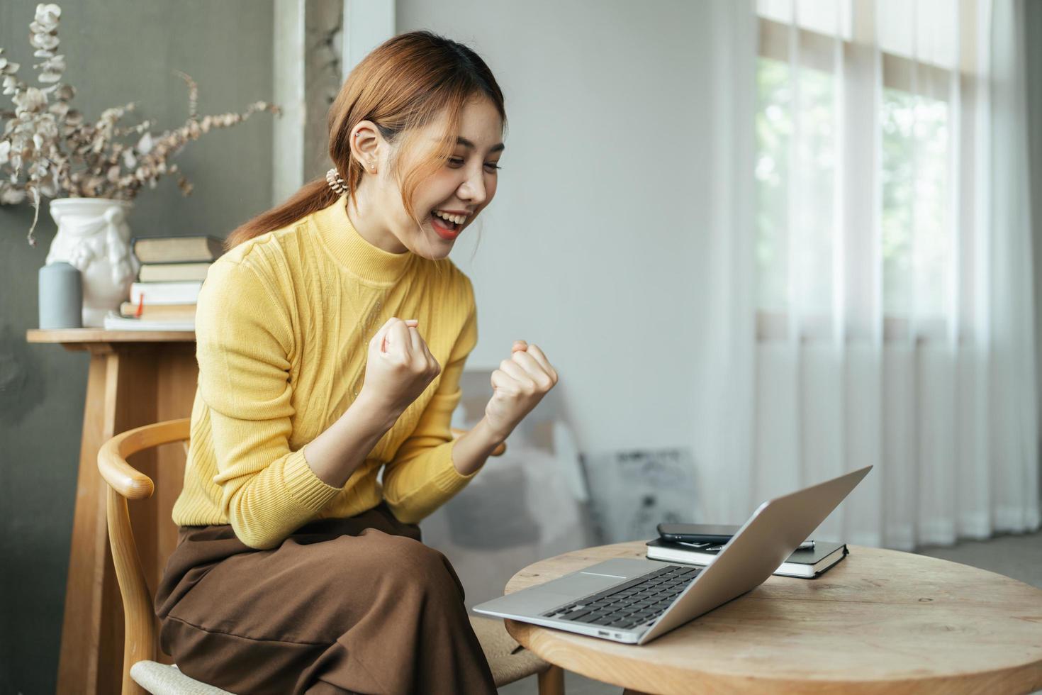 jovem mulher com laptop expressando emoção no escritório em casa, mulher asiática animada se sente eufórica lendo boas notícias online. foto