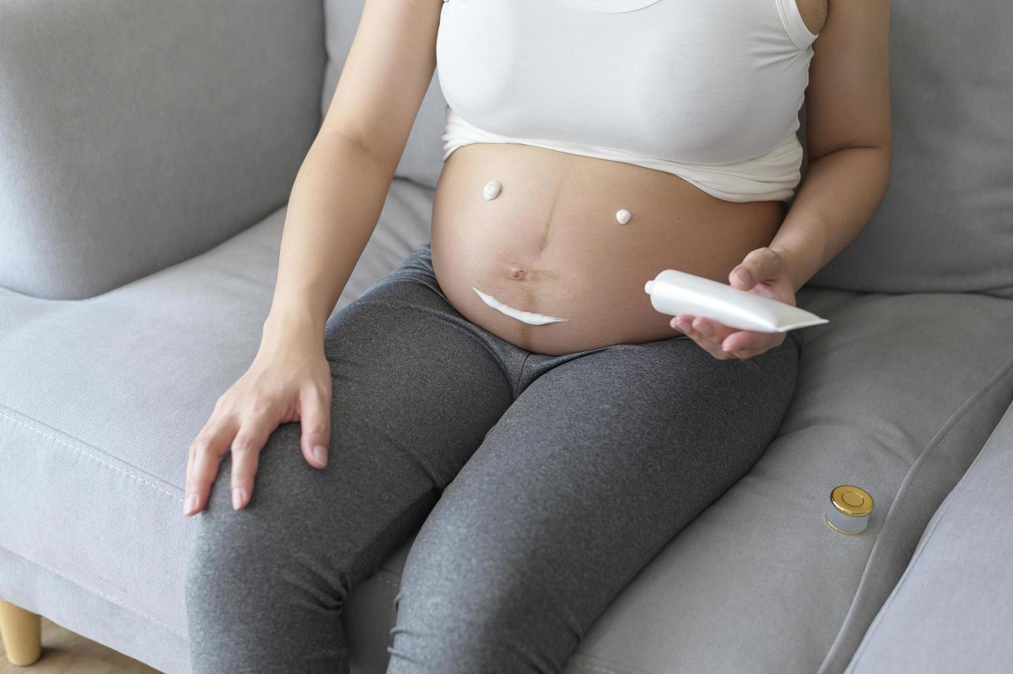 jovem grávida aplicando creme hidratante na barriga, cuidados de saúde e gravidez foto