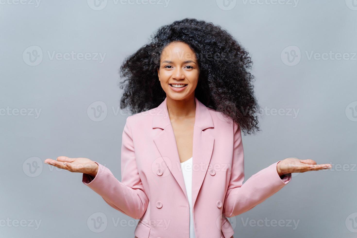tiro isolado de trabalhadora de negócios feminino alegre com penteado afro, veste elegante jaqueta violeta formal, levanta as duas palmas, apresenta algum produto, sorri agradavelmente, isolado sobre fundo cinza. foto
