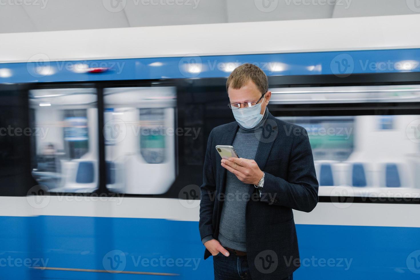 tiro horizontal de trabalhador masculino posa na plataforma do metrô, viaja de transporte público, usa telefone celular moderno para verificar rota, usa máscara de proteção médica contra coronavírus ou gripe. perigo à saúde foto