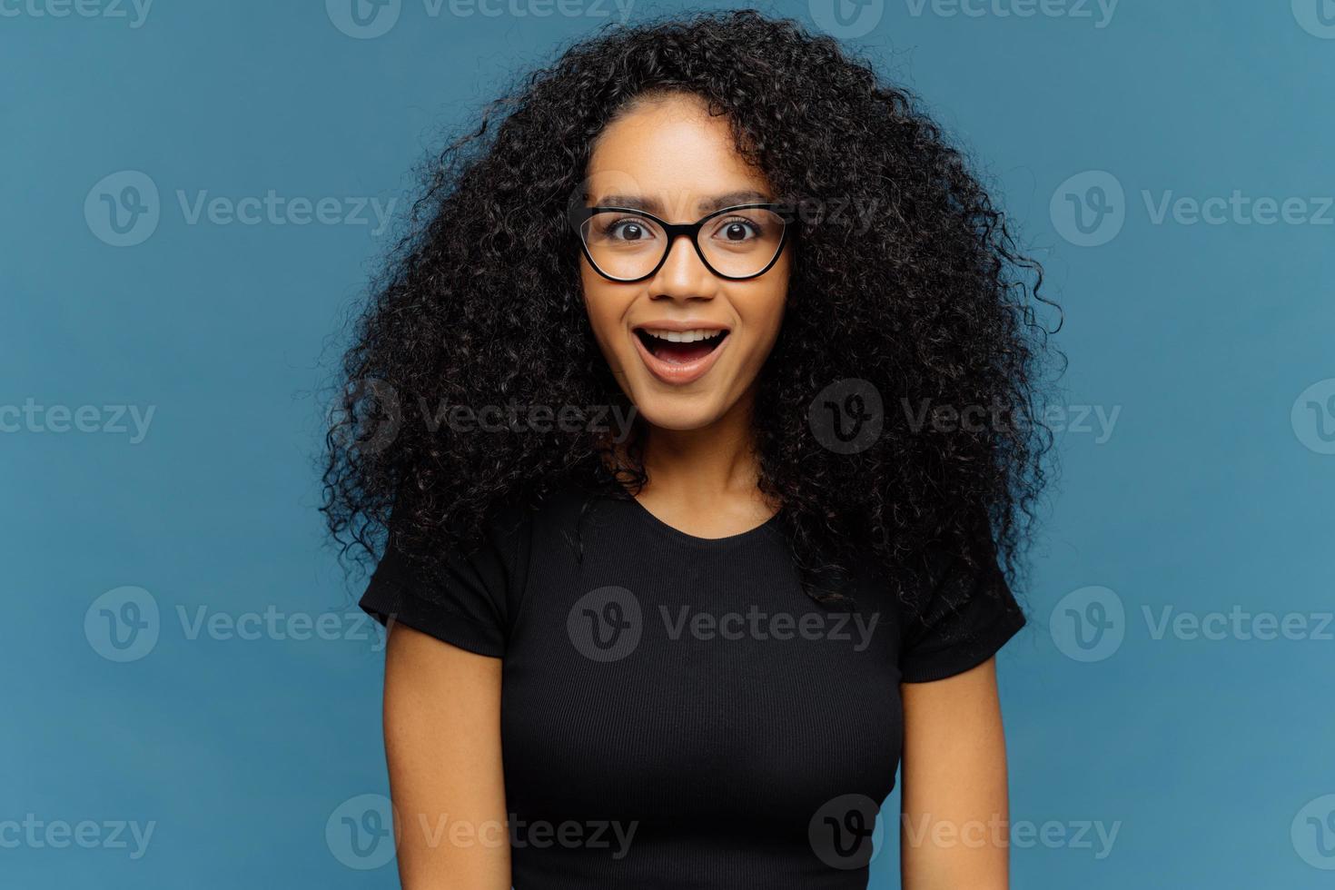 mulher de pele escura agradavelmente surpresa mantém o queixo caído, olha com interesse, tem cabelos cacheados, usa camiseta preta, modelos contra a parede azul. senhora afro-americana olha com choque, prendeu a respiração foto