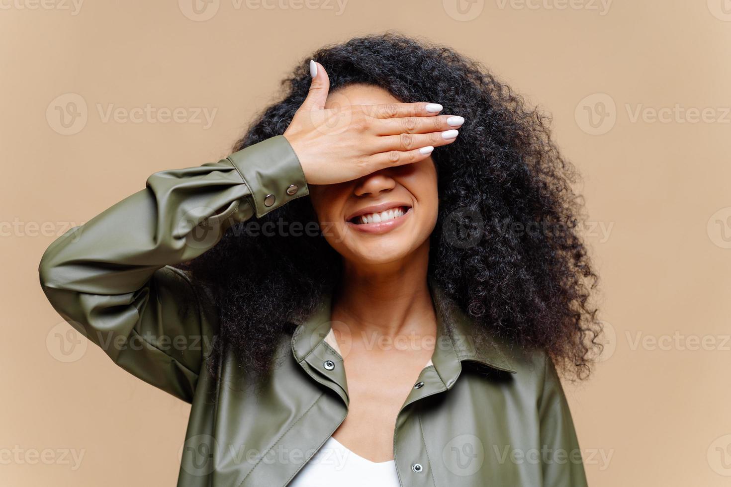 tímida alegre jovem afro cobre os olhos com a palma da mão, tem sorriso, esconde o rosto, tem penteado encaracolado, vestido com roupas elegantes, isolado sobre fundo marrom, espera por surpresa ou presente foto